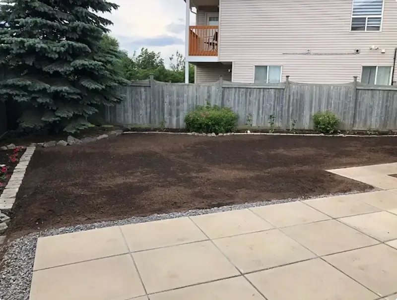 Fresh topsoil grading beside a slab patio and brick-edged flower bed